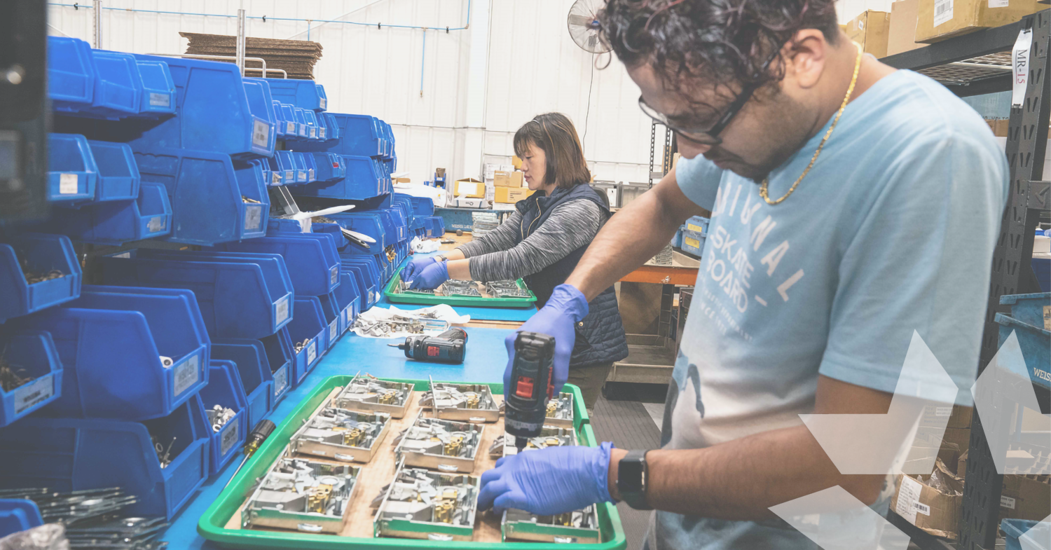 Two PDQ employees assembling lock hardware components on a factory workbench, with bins of parts and a cordless drill in use.