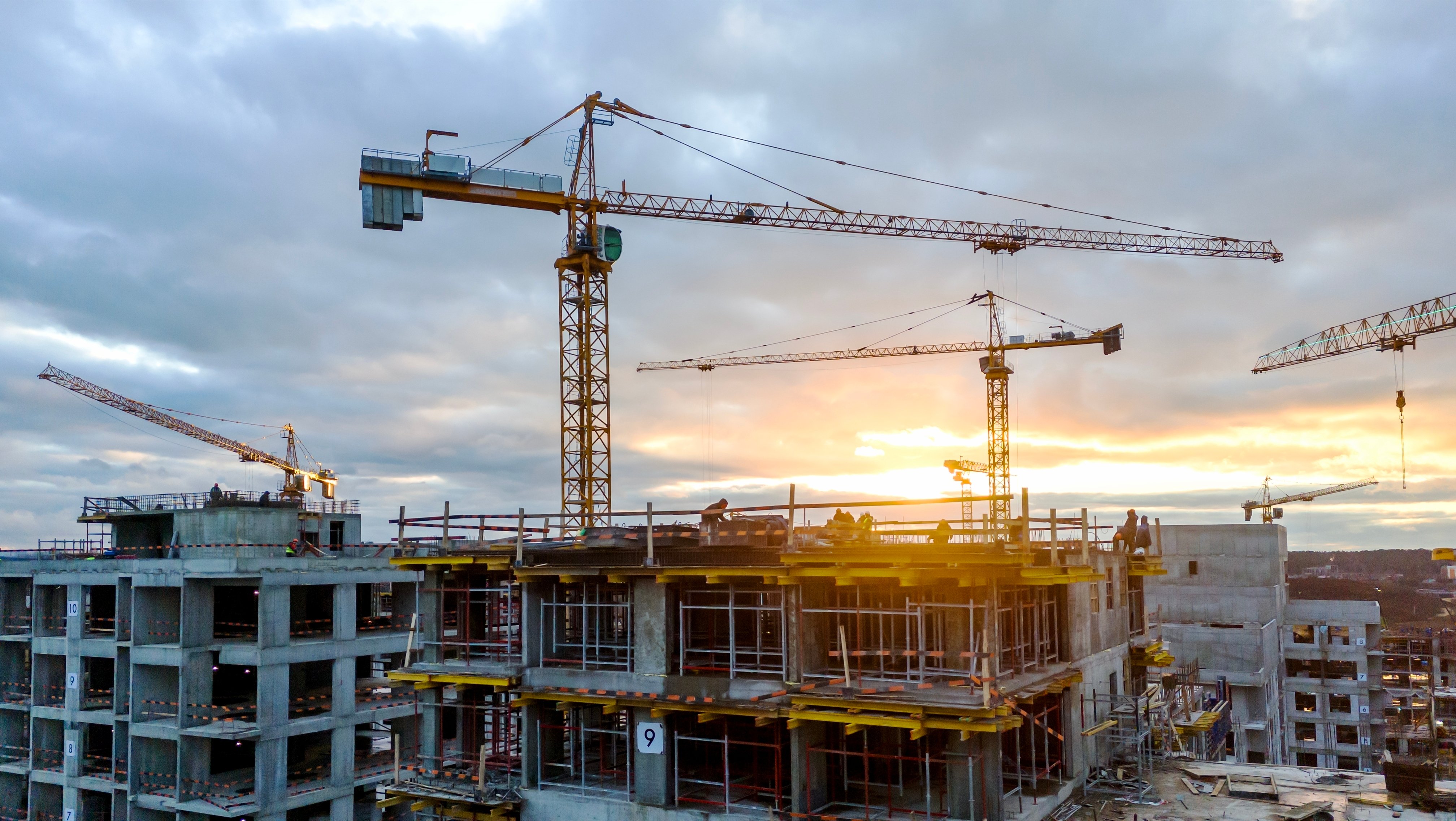 Construction site with multiple tower cranes over a partially built concrete high-rise, silhouetted against a cloudy sunset sky.