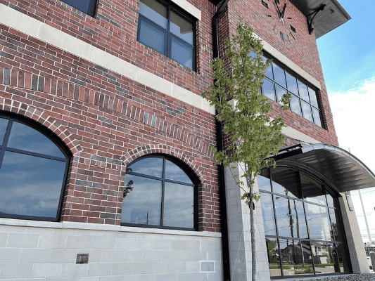 Exterior of the Joseph Koss Public Safety Building, a multi-story brick building with arched and rectangular windows, a clock mounted near the roofline, and a curved metal awning over the entrance, with a young tree in the foreground.