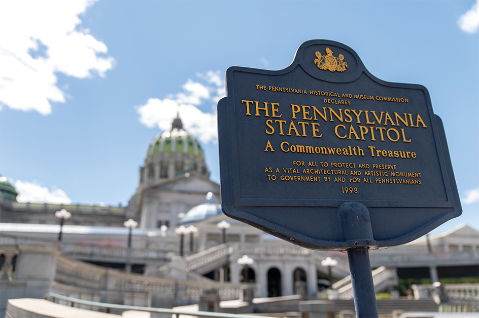 Historical marker declaring the Pennsylvania State Capitol "A Commonwealth Treasure," dated 1998, with the Capitol's green dome and facade visible in the background under a blue sky.