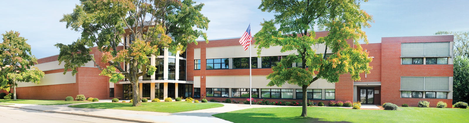Exterior of Racine Lutheran High School with an American flag out front, large windows, landscaped shrubs and flower beds, and mature trees along a curved walkway and lawn under a clear sky.