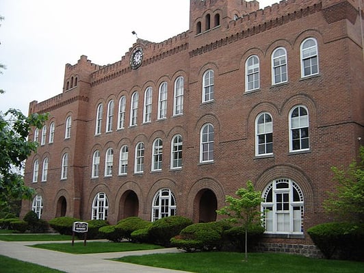 St. Mary's Preparatory School, a historic red brick building with castle-like crenellations, a clock tower, and rows of arched white-trimmed windows, surrounded by trimmed hedges and a lawn under an overcast sky.