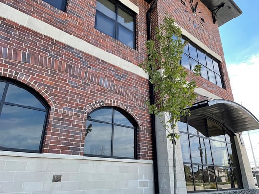 Exterior of the Joseph Koss Public Safety Building, a multi-story brick building with arched and rectangular windows, a clock mounted near the roofline, and a curved metal awning over the entrance, with a young tree in the foreground.