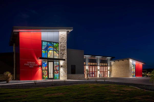 Odessa Fire Rescue Station 9, a modern building with a red panel facade featuring colorful stained-glass artwork, stone accents, and red-framed apparatus bay doors, photographed at dusk with landscape lighting illuminating the entrance.