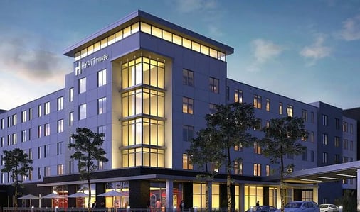 Exterior view of the Hyatt House hotel at dusk, showing a modern multi-story building with a glass-fronted lobby tower lit in warm gold light, illuminated guest room windows, a covered porte-cochère entrance, outdoor patio umbrellas, and landscaped trees against a blue evening sky.