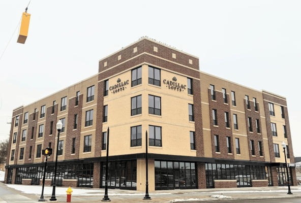 Street-level photo of Cadillac Lofts, a four-story mixed-use brick building on a corner, with tan and brown brick facade, large ground-floor storefront windows including a Jimmy John's, streetlights, and a fire hydrant in the foreground on a snowy day.