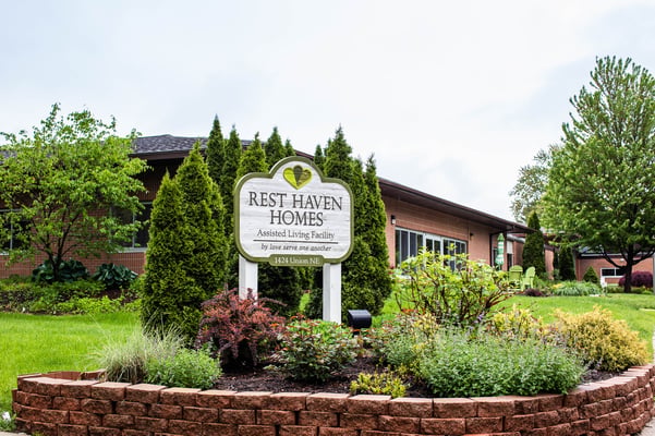 Photo of the Rest Haven Homes assisted living facility wooden sign with a heart logo reading "by love serve one another," set among landscaped shrubs and evergreen trees in front of a brick single-story building.