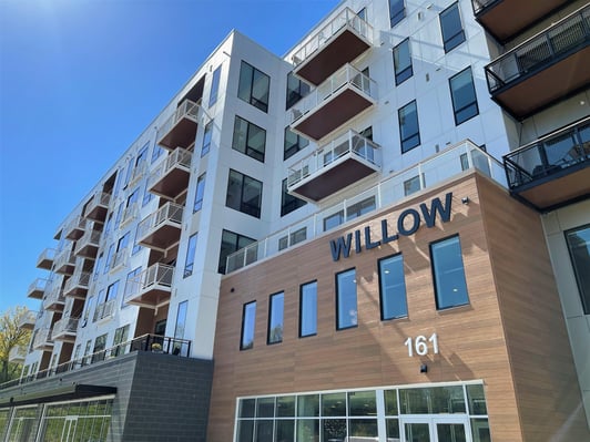 Upward-angled photo of the Willow apartment building at 161 Riverheath, showing a modern multi-story facade with white paneling, wood-tone siding, staggered balconies with cable railings, and large glass storefront entrance under a clear blue sky.