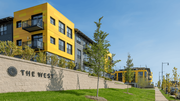 Photo of The West apartment complex, featuring a stone entrance wall with logo signage on a grassy slope, leading up to modern multi-story buildings with yellow and gray panel facades, black balcony railings, young trees, and a sidewalk along the street.