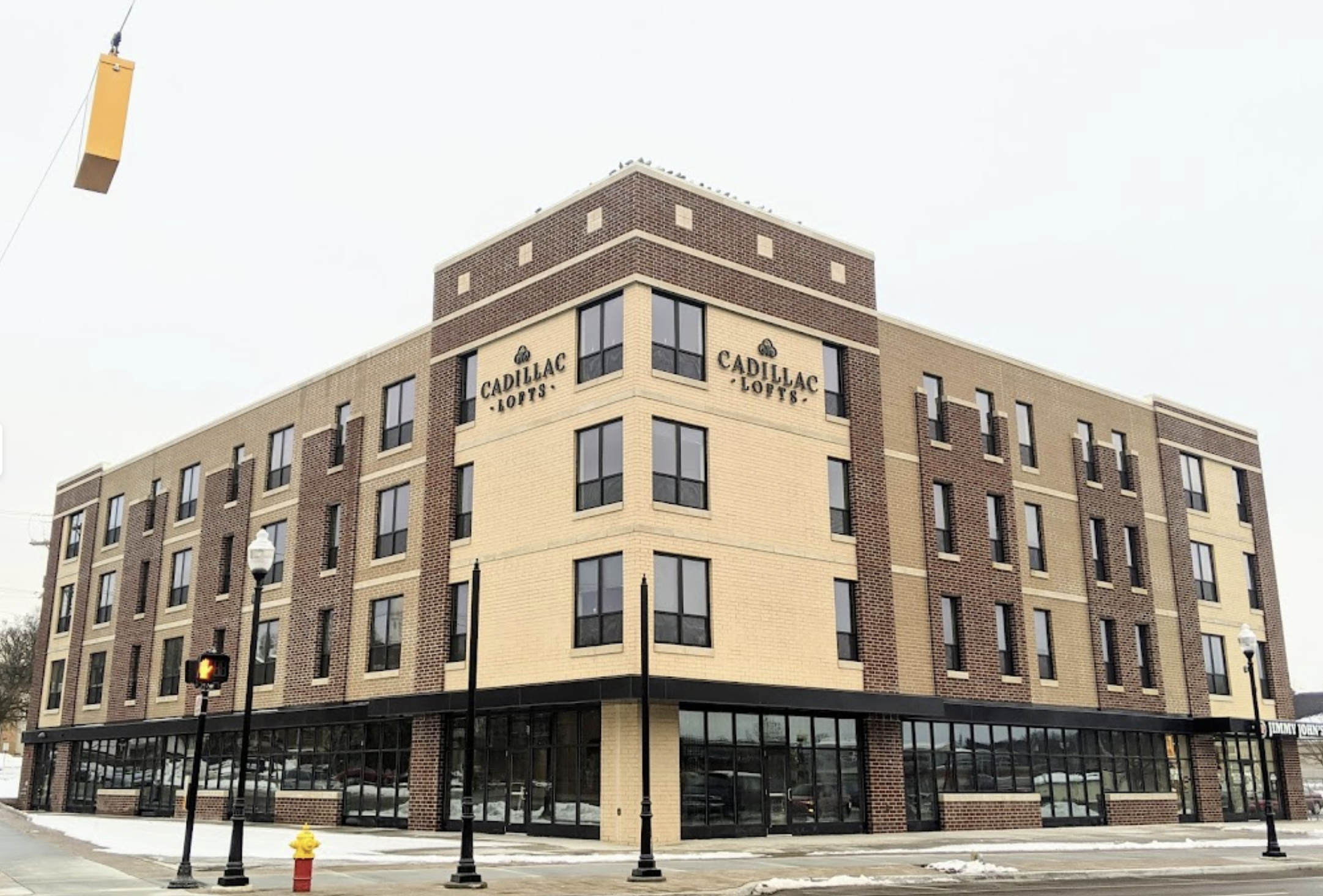 Street-level photo of Cadillac Lofts, a four-story mixed-use brick building on a corner, with tan and brown brick facade, large ground-floor storefront windows including a Jimmy John's, streetlights, and a fire hydrant in the foreground on a snowy day.