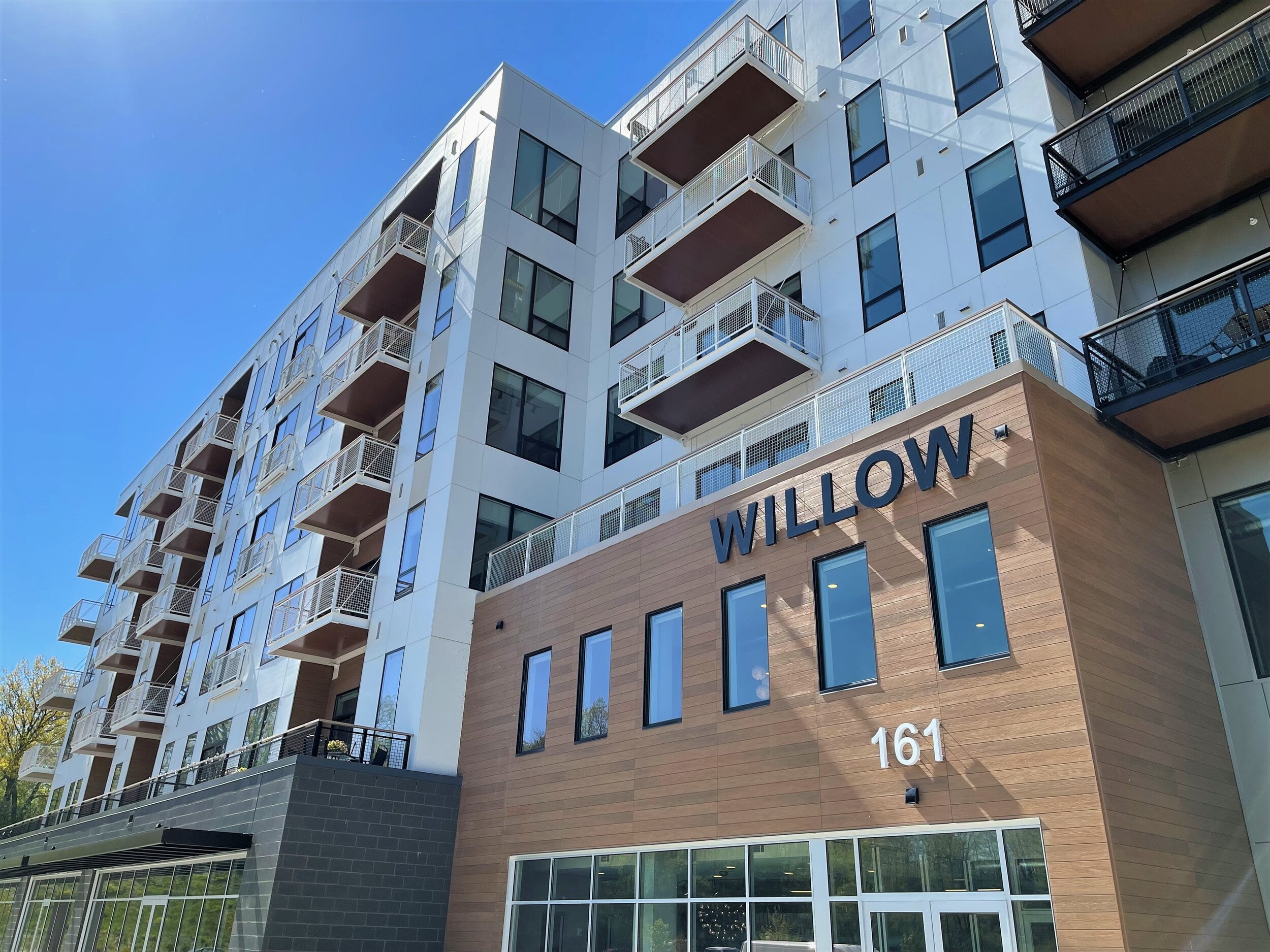 Upward-angled photo of the Willow apartment building at 161 Riverheath, showing a modern multi-story facade with white paneling, wood-tone siding, staggered balconies with cable railings, and large glass storefront entrance under a clear blue sky.